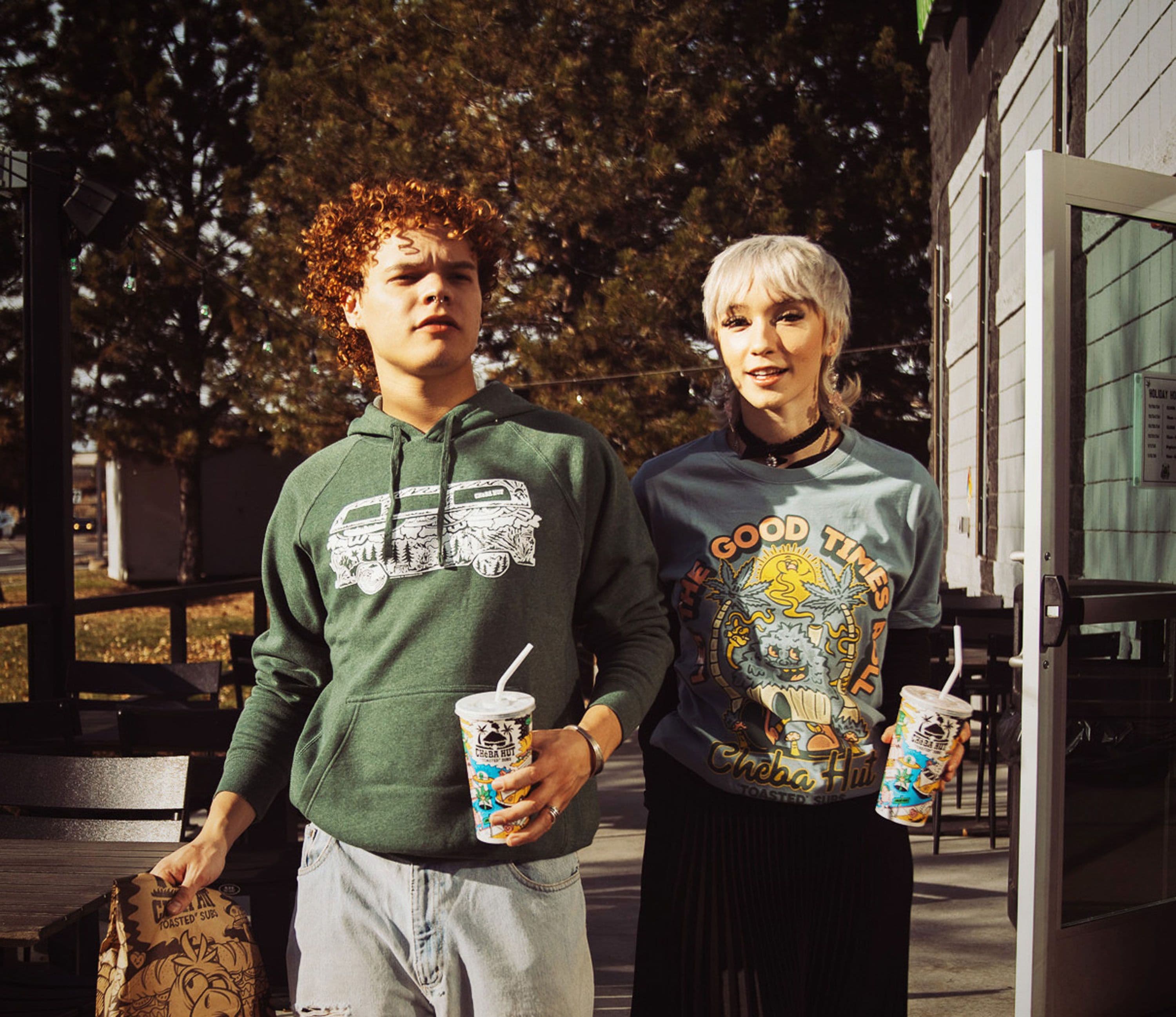 Two people wearing Cheba Hut merchandise and holding drinks outside a shop
